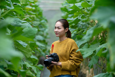 Young woman holding camera while standing amidst plants