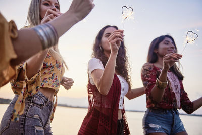 Friends holding sparklers while standing against sky