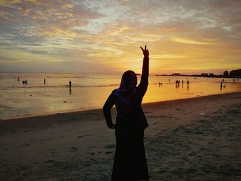 Rear view of silhouette young woman with hand raised standing at beach against cloudy sky during sunset