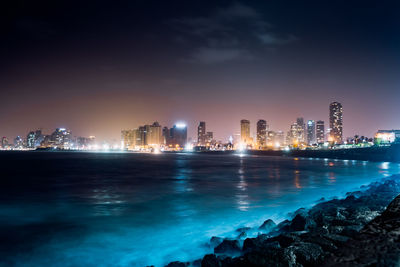 Illuminated buildings by sea against sky at night