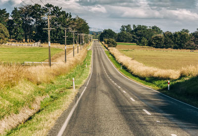 Empty road amidst trees on field against sky