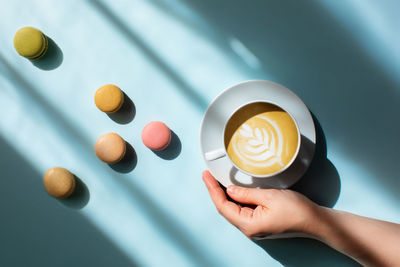 Morning coffee in a woman's hand with delicious macaroons scattered around in the sunlight.