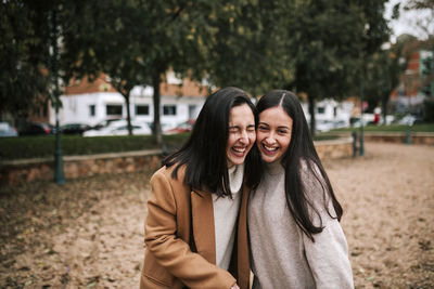 Cheerful sisters standing against trees in park during winter