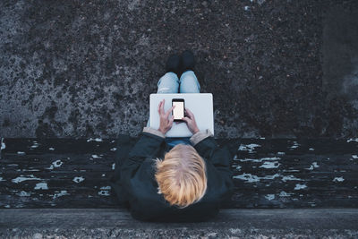 Directly above shot of woman with laptop using mobile phone while sitting on bench against retaining wall