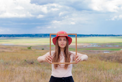 Girl in pink straw hat holds empty wooden picture frame  nature landscape portrait. creative idea.