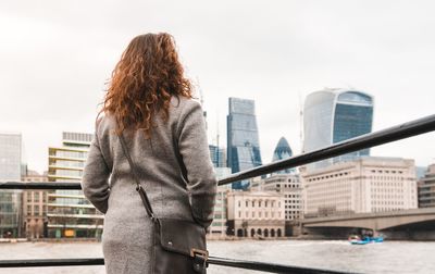 Rear view of woman standing by railing against river and buildings in city