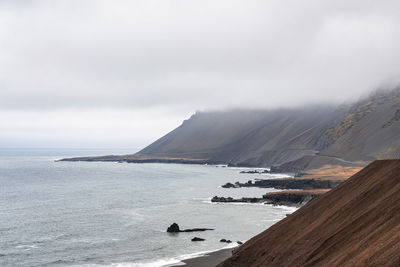 Beautiful view of steep brown slope by beach against cloudy sky in foggy weather
