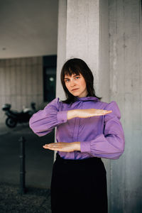 Portrait of young woman gesturing equal sign while standing against wall