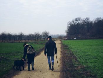 Rear view of men walking with dog on road amidst field against sky