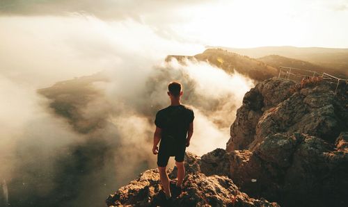 Rear view of man standing on rock during foggy weather against sky