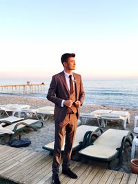 Young man looking away while standing at beach in suit against sky