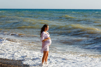 Portrait beautiful young pregnant woman, against blue sea on beach, thoughtful