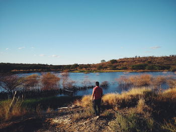 Rear view of man standing on field by lake against clear sky