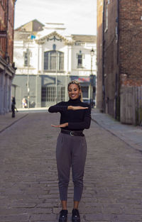 Full length portrait of woman gesturing while standing on footpath against buildings