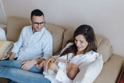 Happy mother holding newborn baby son in sofa with father looking