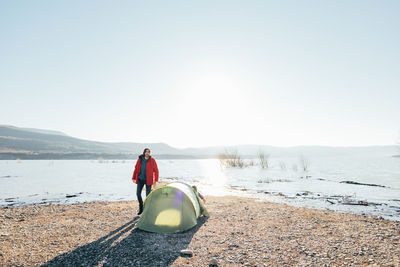 Male explorer in outerwear standing near tent at campsite near calm pond in highlands on sunny autumn day