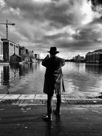 Rear view of mid adult man standing by river on footpath against cloudy sky in city