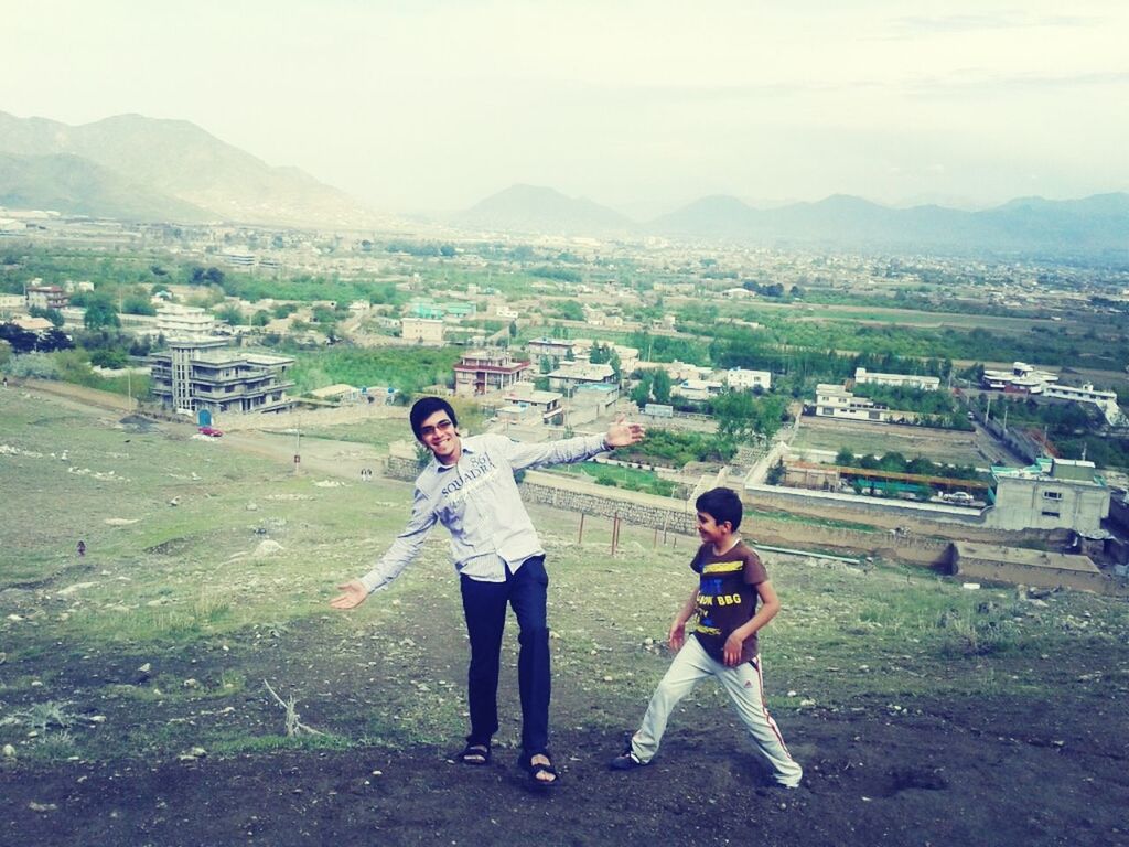 lifestyles, casual clothing, person, leisure activity, full length, young adult, smiling, happiness, young men, field, sitting, sky, looking at camera, mountain, togetherness, standing