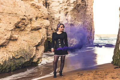 Young woman with purple distress flare standing by rock formations at beach