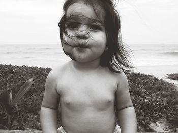 Shirtless baby girl sitting by sea against sky