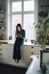 Full length of smiling girl using mobile phone while sitting on kitchen counter