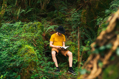 Caucasian man in a yellow t-shirt and a black hat sitting in the middle of the jungle reading a book