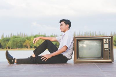 Side view of smiling teenage boy sitting by television set against sky