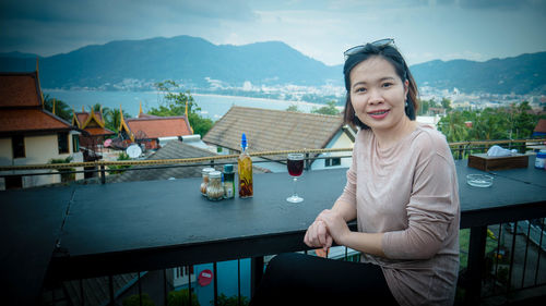 Portrait of smiling woman with drink on table sitting in cafe against mountains