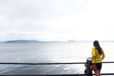 Rear view of mid adult woman looking at sea while standing by railing against cloudy sky