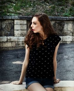 Young woman looking away sitting on retaining wall against street