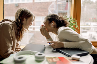 Side view of teenage female student gossiping with friend sitting in high school