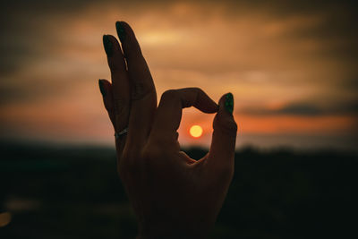 Sun seen through cropped hand against cloudy sky during sunset