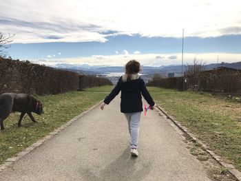 Rear view of child walking on footpath amidst field against sky
