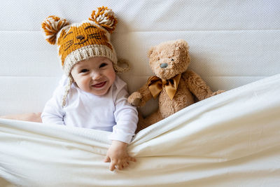 Happy boy with wool cap lying with teddy bear under the blanket on bed