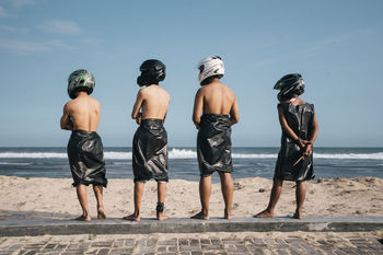 REAR VIEW OF MEN STANDING AT BEACH AGAINST SKY