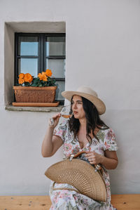 Beautiful young woman sitting on bench in front of old house, drinking wine