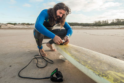 Man tying rope in surfboard at beach against sky