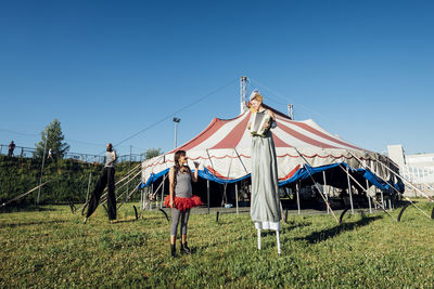 Female acrobat looking at performer playing accordion while standing with stilts on meadow