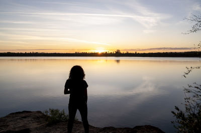 Rear view of woman standing at lakeshore during sunset