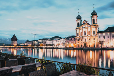 Illuminated buildings by river against sky in city at dusk