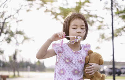 Girl blowing bubbles while holding teddy bear in park