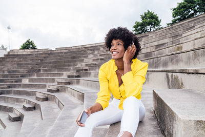Smiling african american female with afro hairstyle sitting on stairs in city and using a cellphone