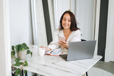 Smiling woman with long hair in white cardigan working on laptop using mobile phone in office