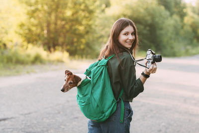 A charming young girl walks in nature, holding a backpack on her shoulder, from which her dog 