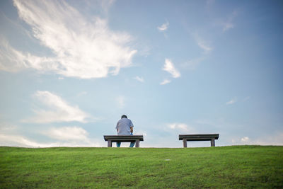 Rear view of man sitting on bench at grassy field against blue sky