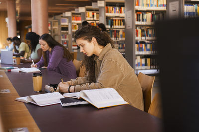 Young female student writing in book while sitting at desk studying inside university library