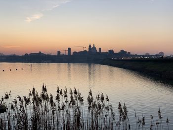 Silhouette buildings by lake against sky during sunset