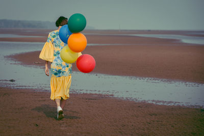 Rear view full length of young woman holding colorful balloons at beach