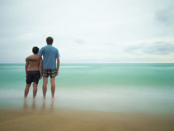 Rear view of man with son standing at beach against cloudy sky