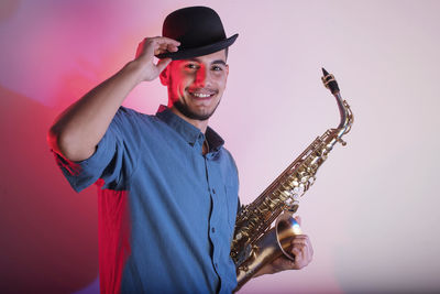 Portrait of young man wearing hat holding saxophone while against wall